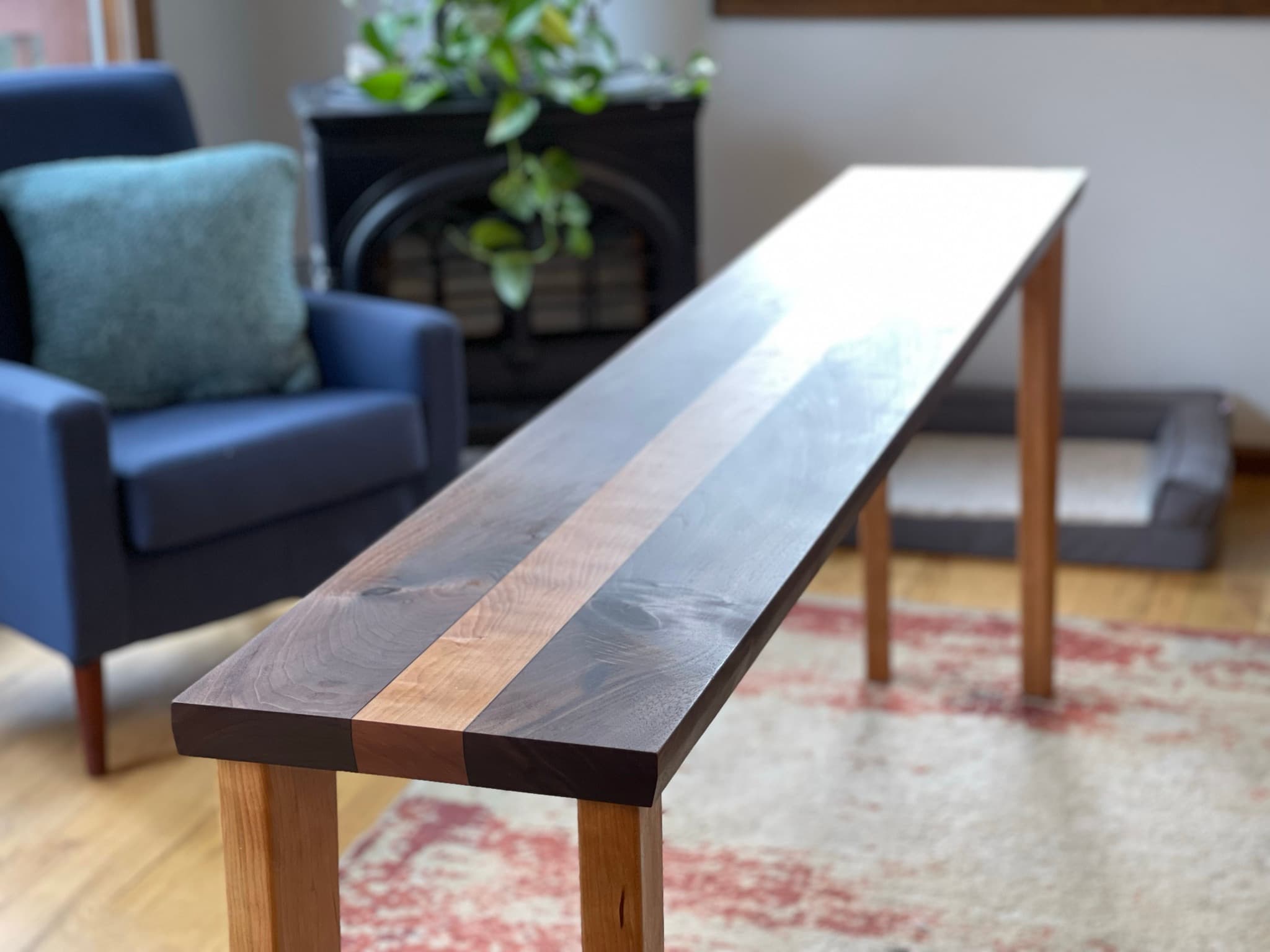 Long, narrow walnut-and-maple console table installed in a living room, blue chair and fireplace in the background, late afternoon light raking across the top.