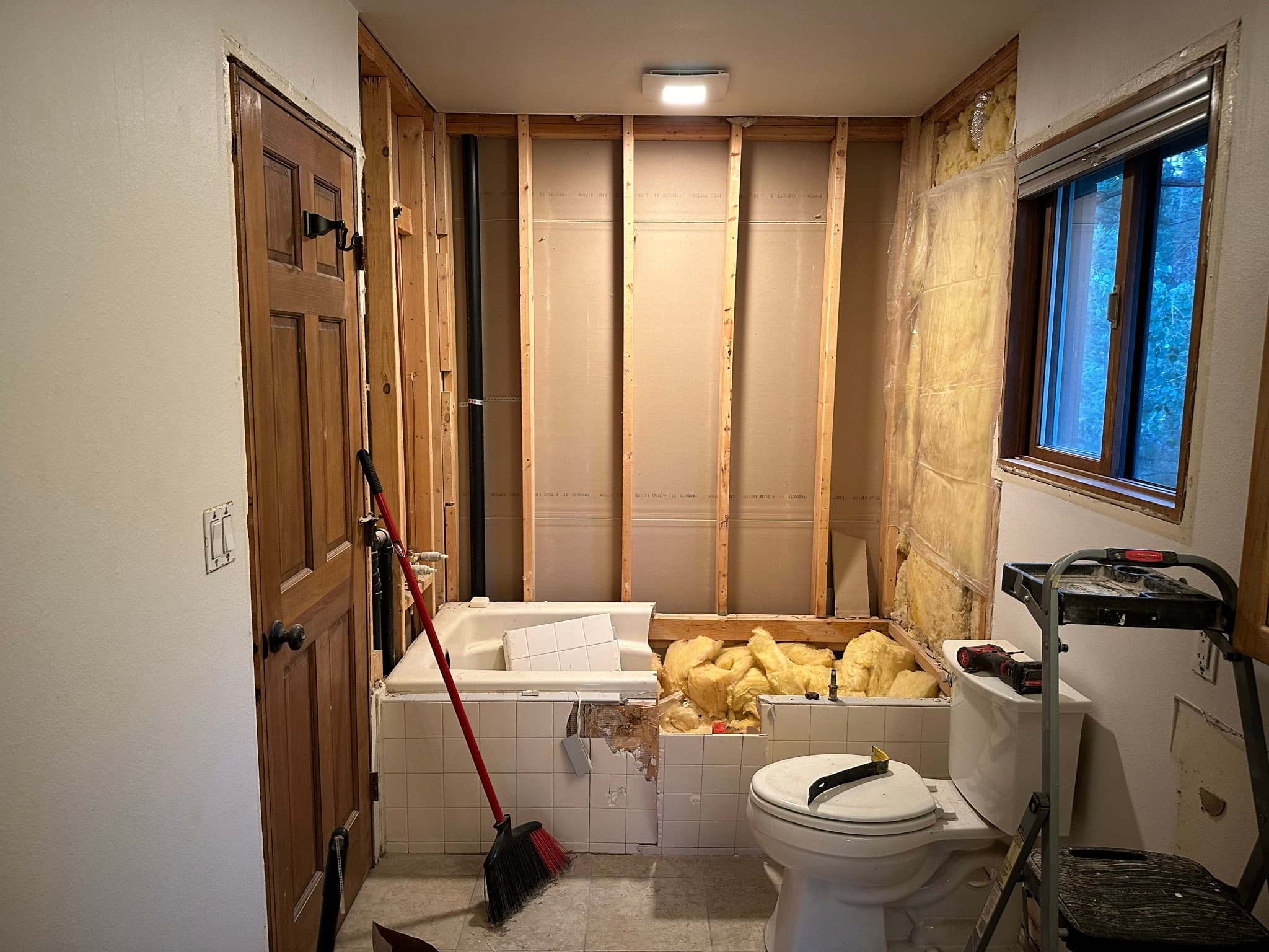 Mid-demo: wall behind the tub stripped to studs, fiberglass insulation exposed, old ABS waste pipe visible, some tile still clinging to the tub surround.