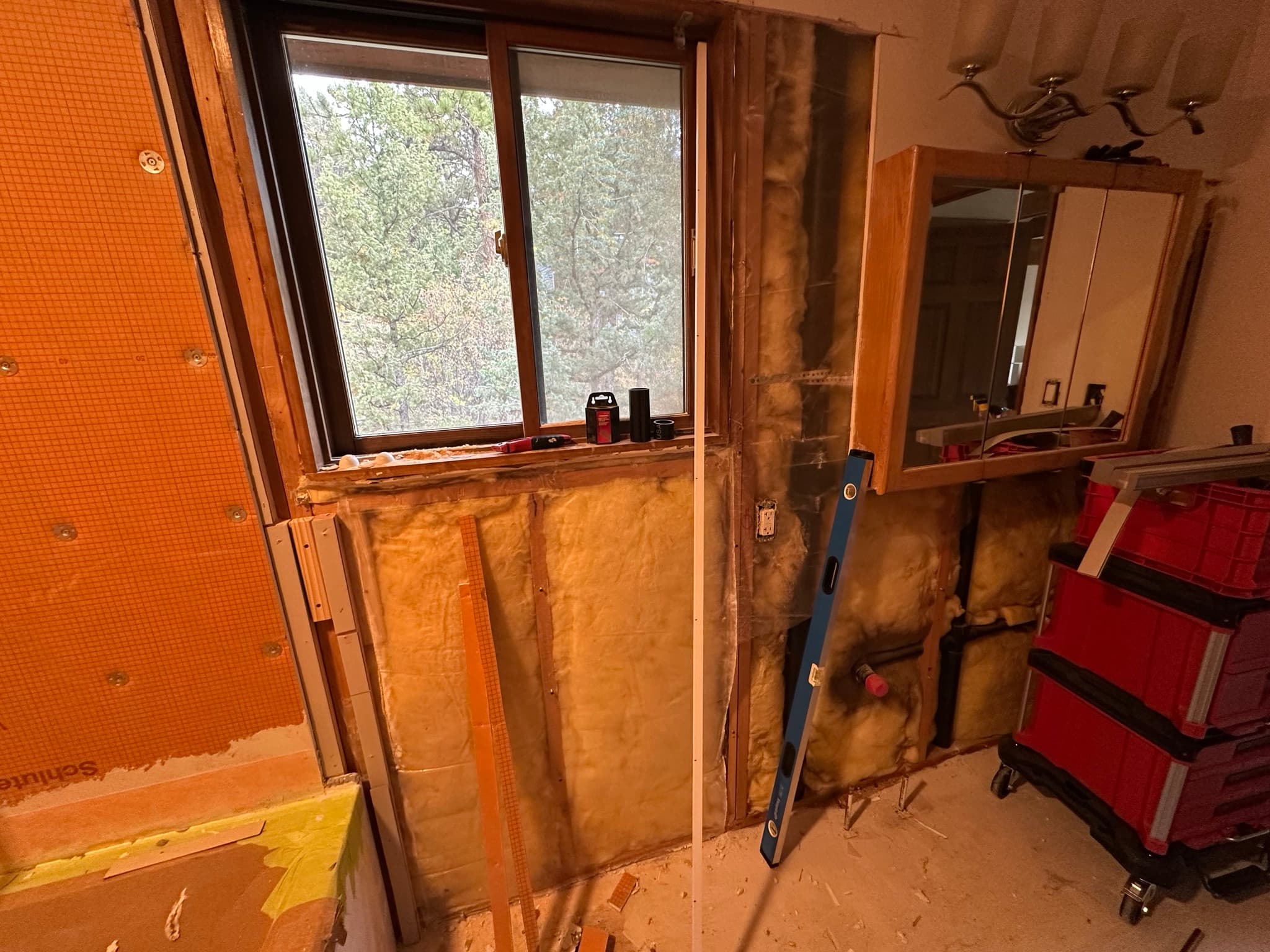 Vanity wall stripped: fiberglass insulation around the window opening, level leaning against the studs, Milwaukee Packout containers on the floor.