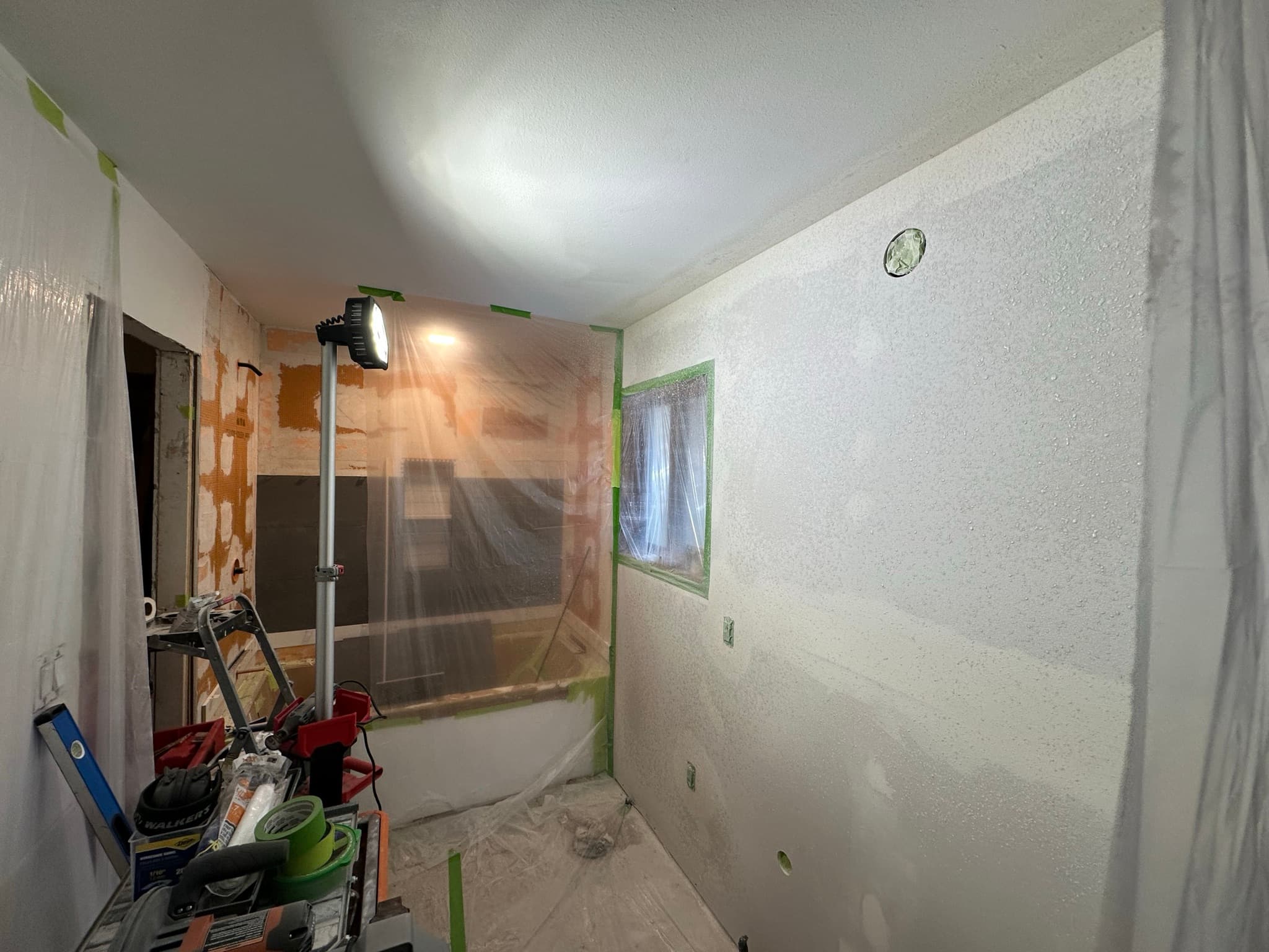 Dry walls re-finished with knockdown texture, freshly sprayed and drying; plastic sheeting and painter's tape masking the tile and tub area to protect them.