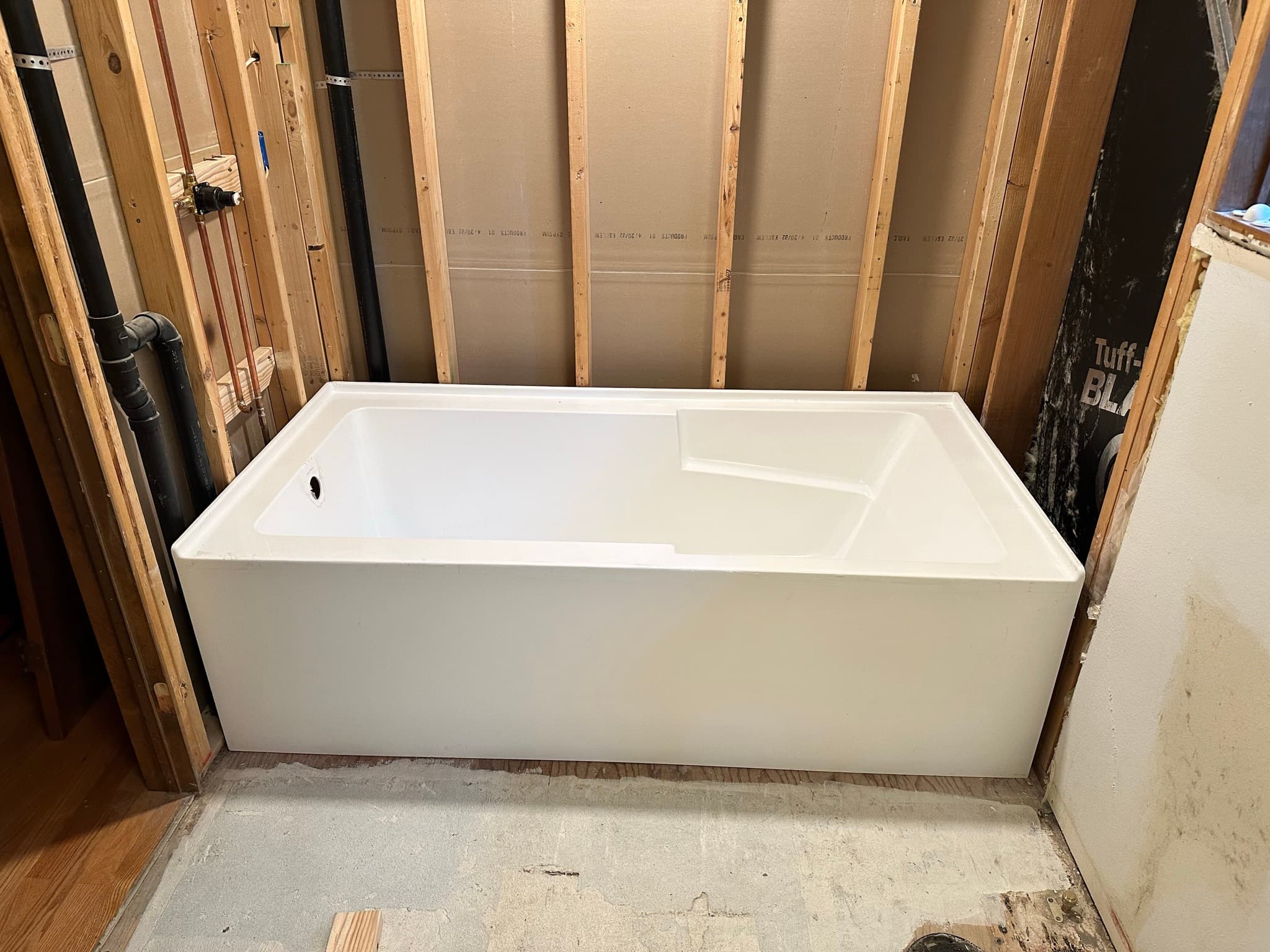 New alcove tub installed in the rough opening, walls still open to studs, plumbing rough-in visible behind.