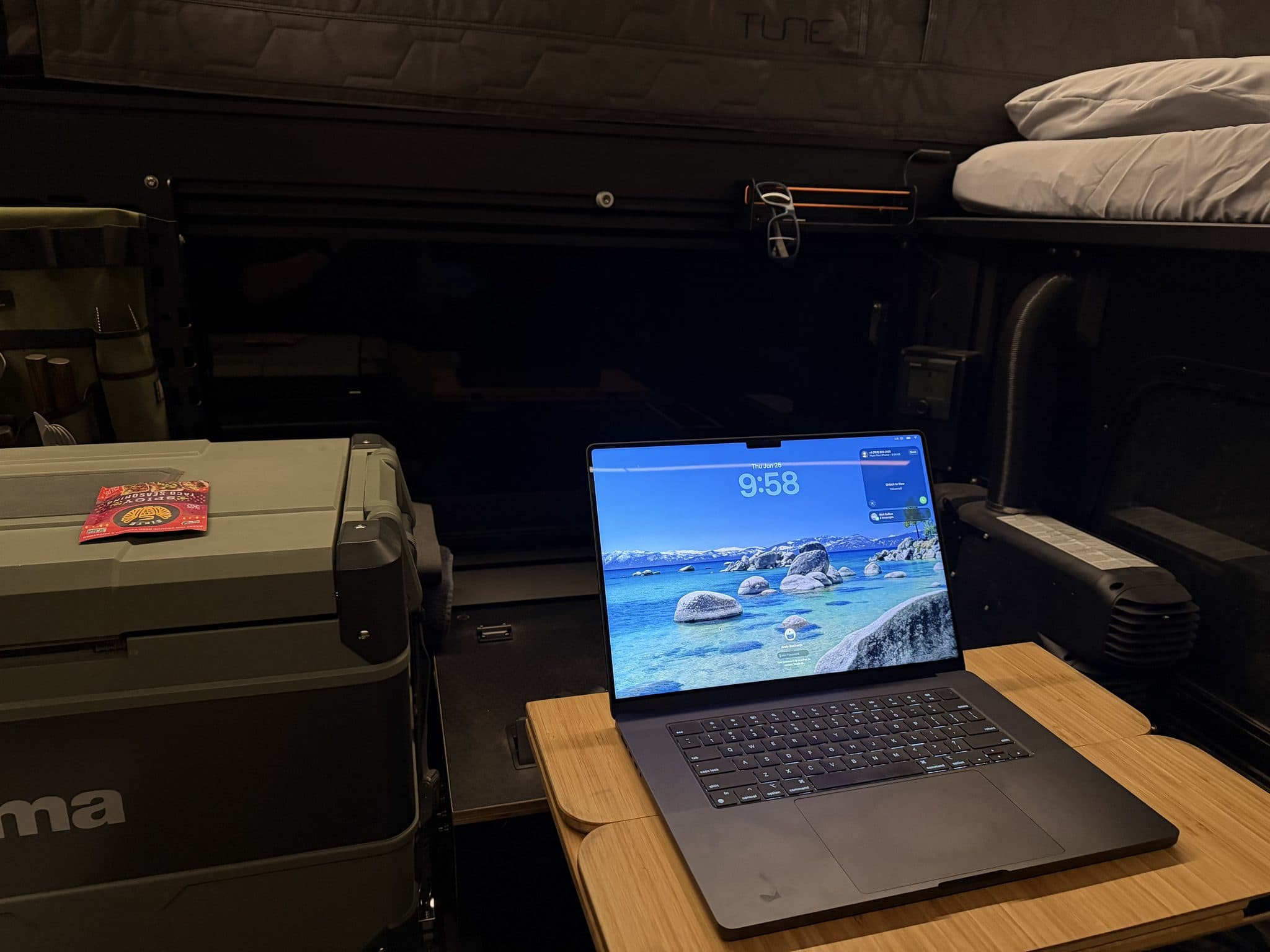 Camper interior at night: laptop open on a bamboo table, fridge at left, made bed at right — everything running off the house battery.